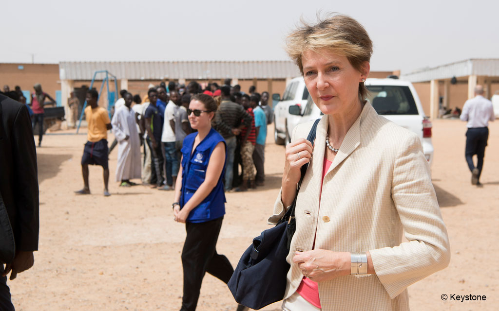 La conseilère fédérale Simonetta Sommruga visite le centre d’accueil et de transit pour migrants de l’OIM à Agadez. À l’arrière-plan, on voit des migrants et des baraquements du camp.