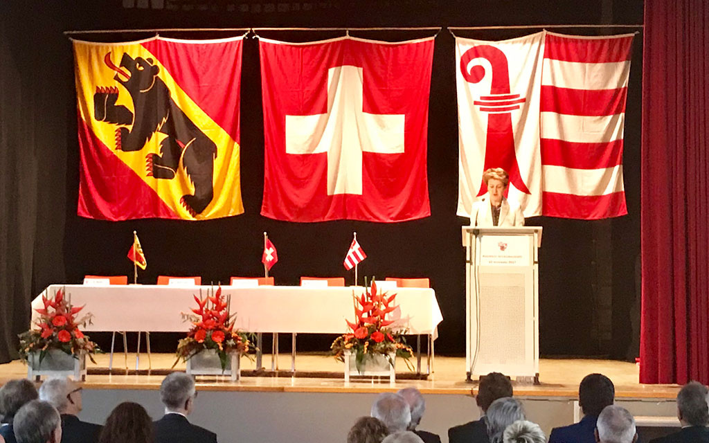 La conseillère fédérale Simonetta Sommaruga est debout sur l’estrade au pupitre de l’orateur, derrière elle les drapeaux du canton de Berne, de la Suisse et du canton du Jura.